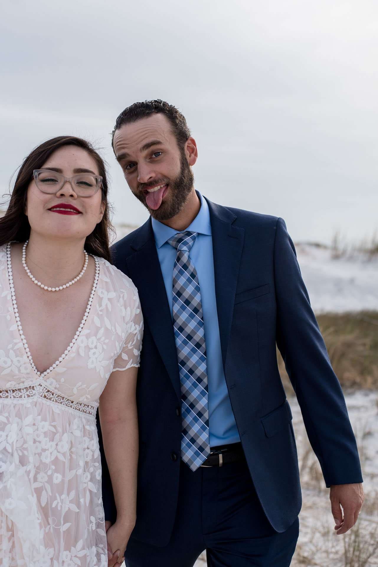 Smiling woman in white lace dress and pearls beside a bearded man in a navy suit sticking out his tongue, snowy dunes behind