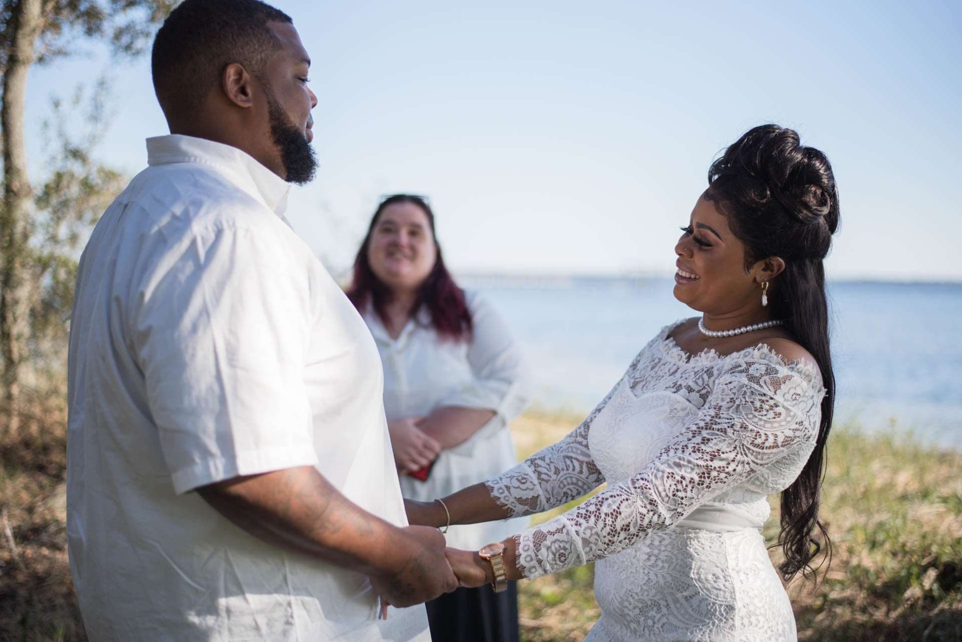 Couple holding hands and smiling during an outdoor waterfront ceremony, bride in a white lace gown, officiant behind them