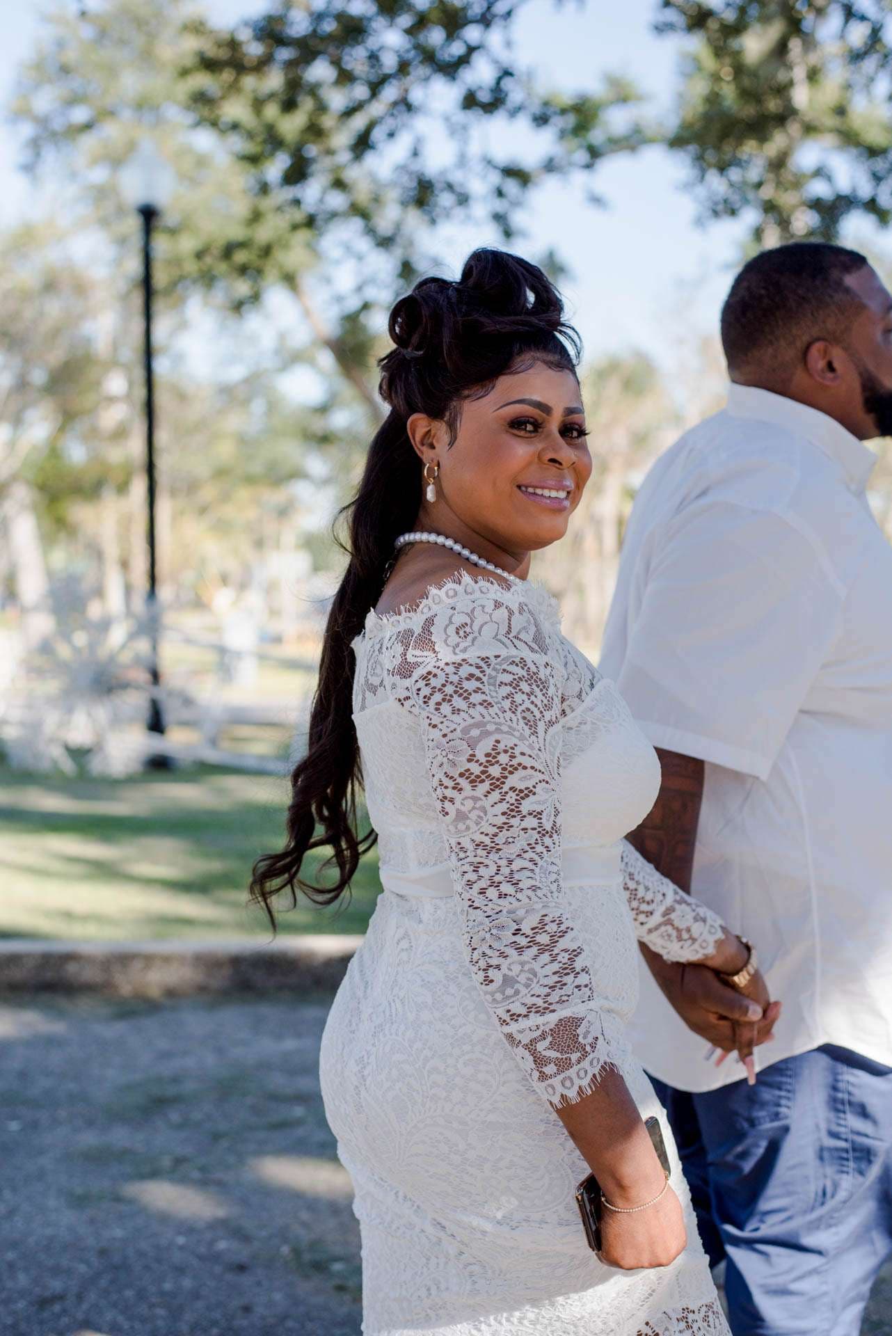 Smiling woman in a white lace dress with a high ponytail looks back while holding hands with a man in a white shirt