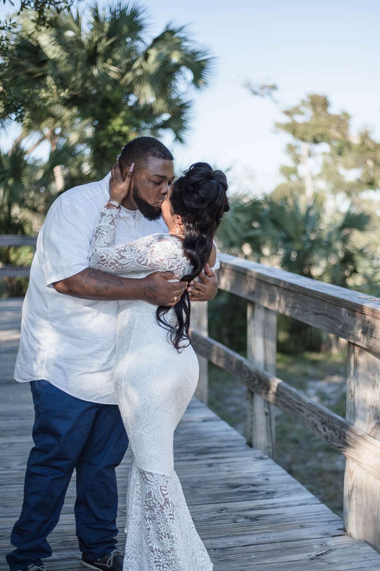 Couple embracing and kissing on a wooden boardwalk, woman in a white lace gown, palm trees behind them