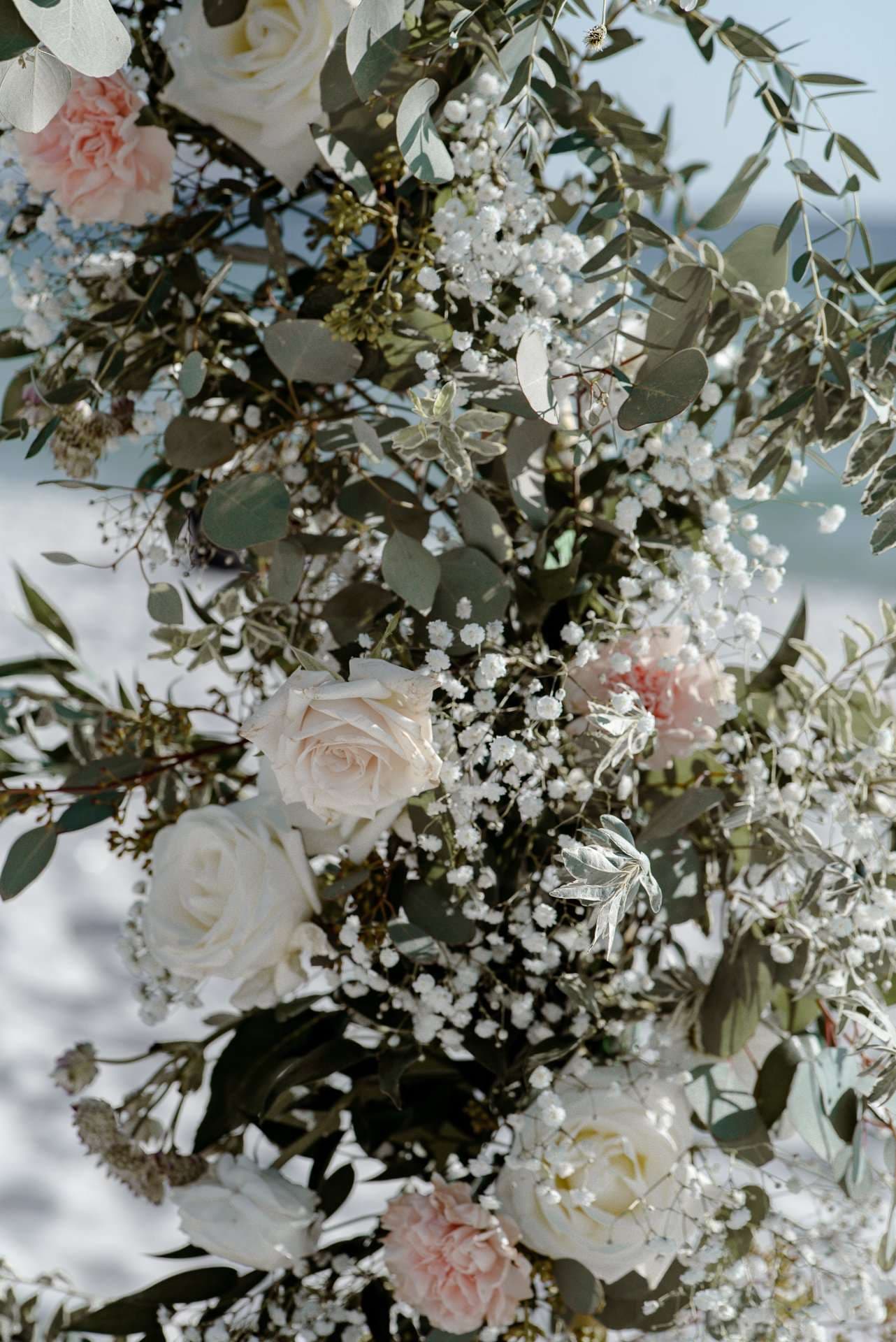 Wedding floral arrangement of white and blush roses with baby's breath and eucalyptus against a soft blurred seaside backdrop