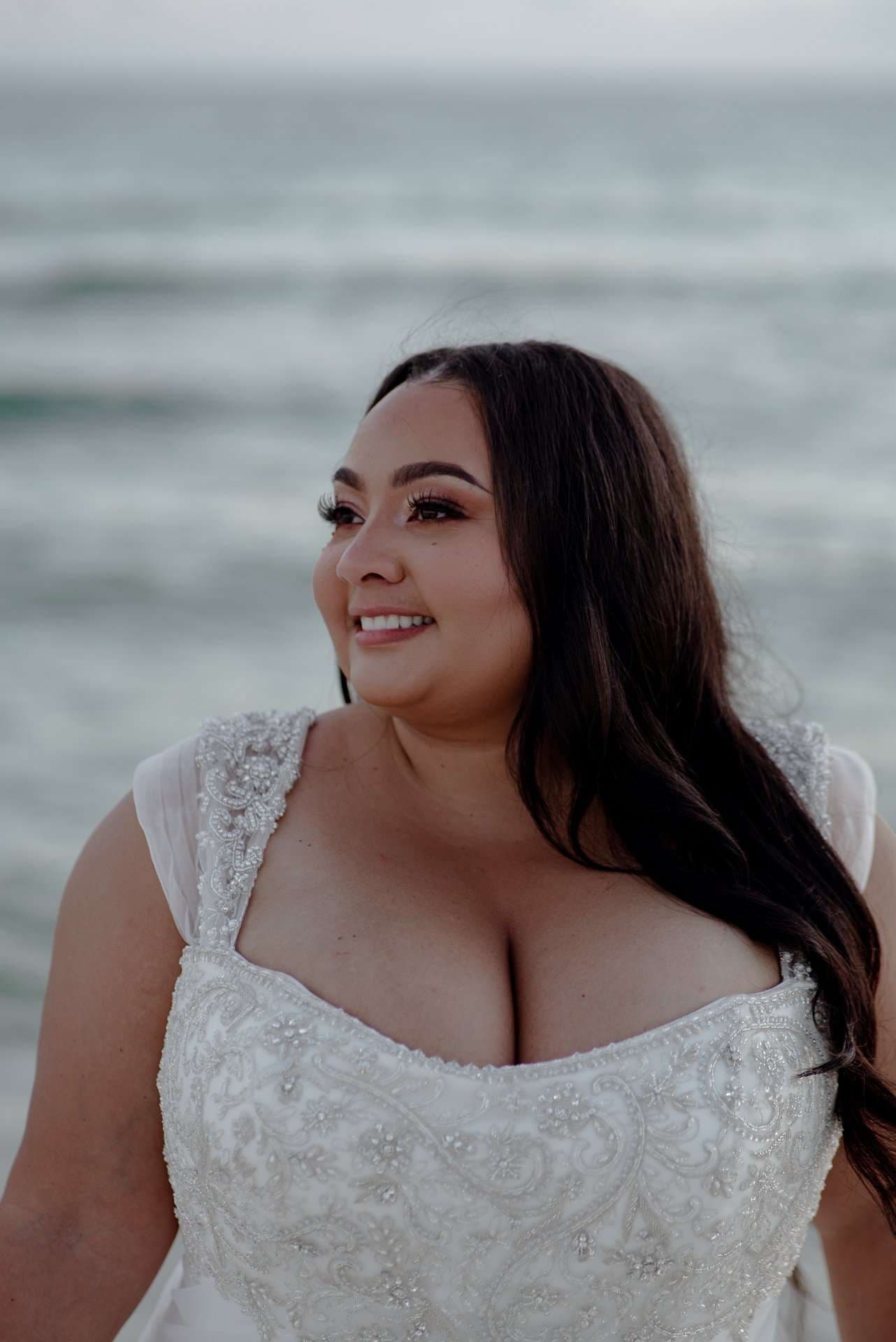 Smiling bride in a beaded white lace gown with long dark wavy hair, looking off to the side by the ocean
