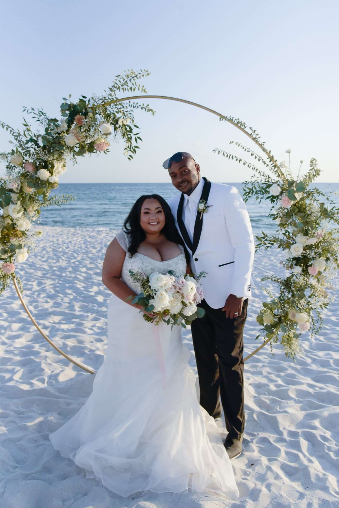 Couple posing in front of a round floral arch on a white sand beach, bride in a white gown holding a bouquet