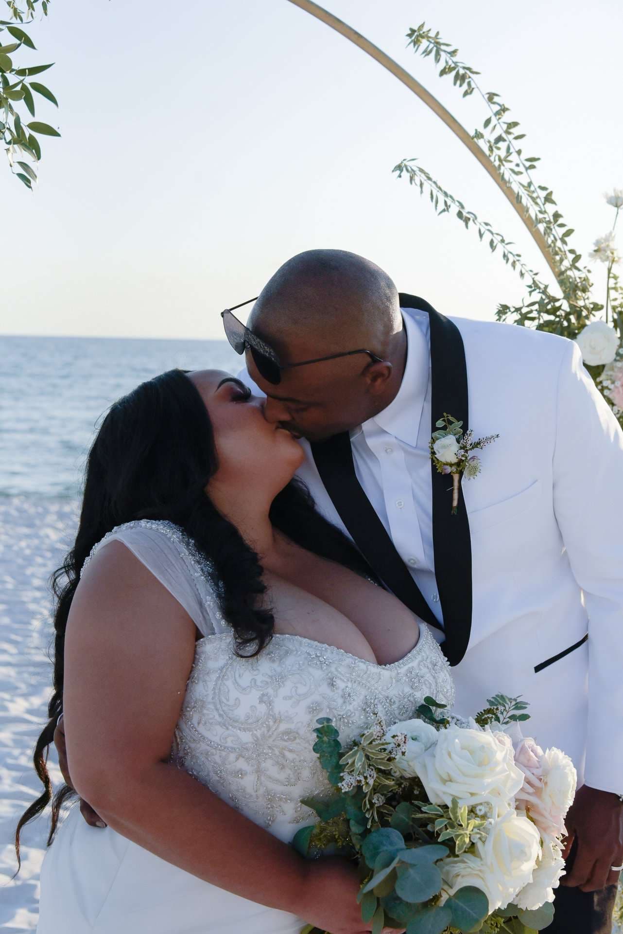 Couple kissing beneath a round floral arch on the beach, she in a beaded white gown, he in a white tuxedo jacket