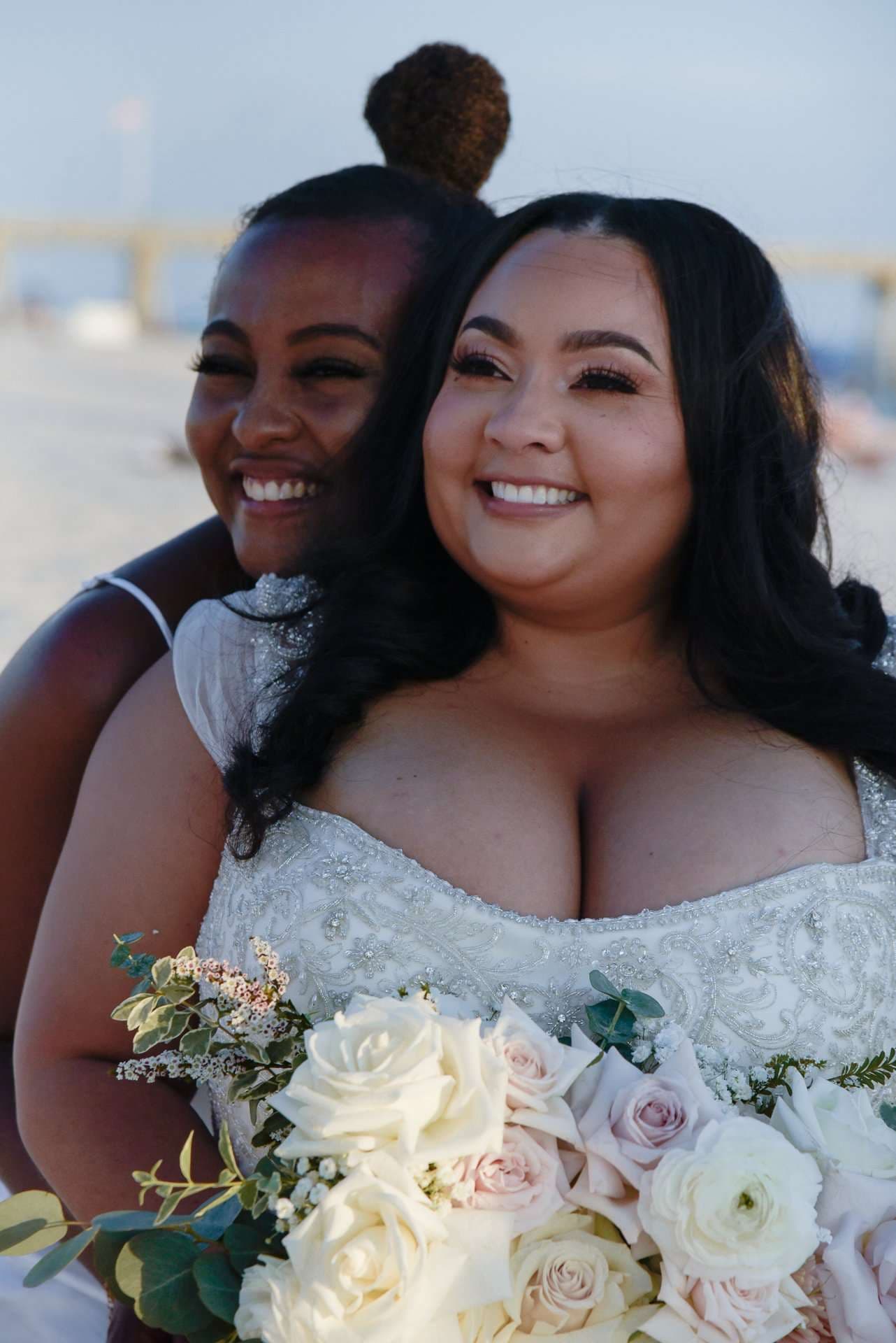 Smiling bride in a beaded white gown holding a pink and white rose bouquet, a laughing woman hugging her from behind