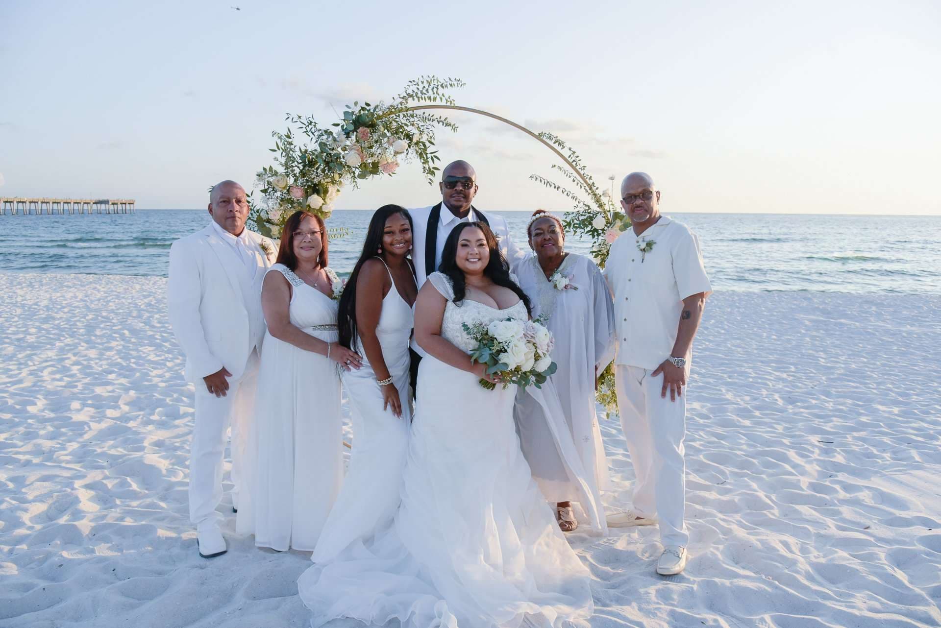 Wedding party in white posing around a floral arch on a white sand beach with the sea behind