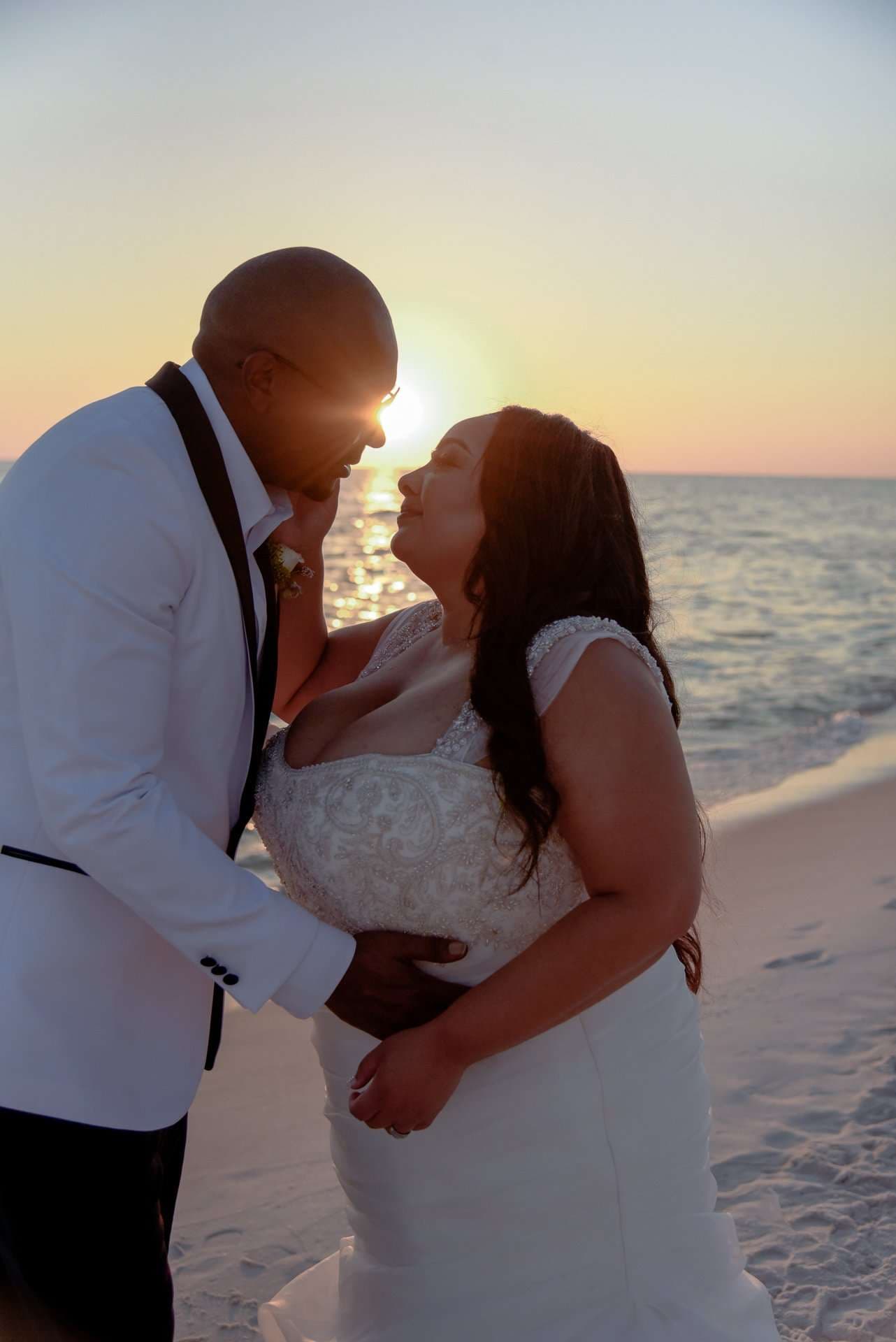 Couple in wedding attire share a tender moment on the beach at sunset, he in a white tuxedo jacket, she in a beaded gown