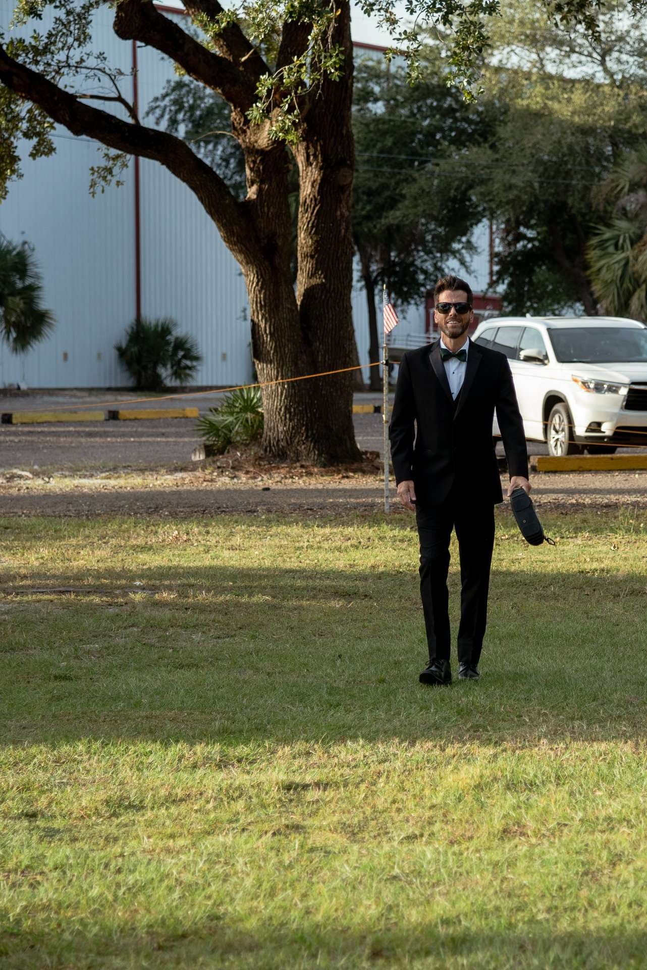 Man in a black tuxedo, bow tie and sunglasses walking across a grassy lawn holding a shoe beneath a large oak tree