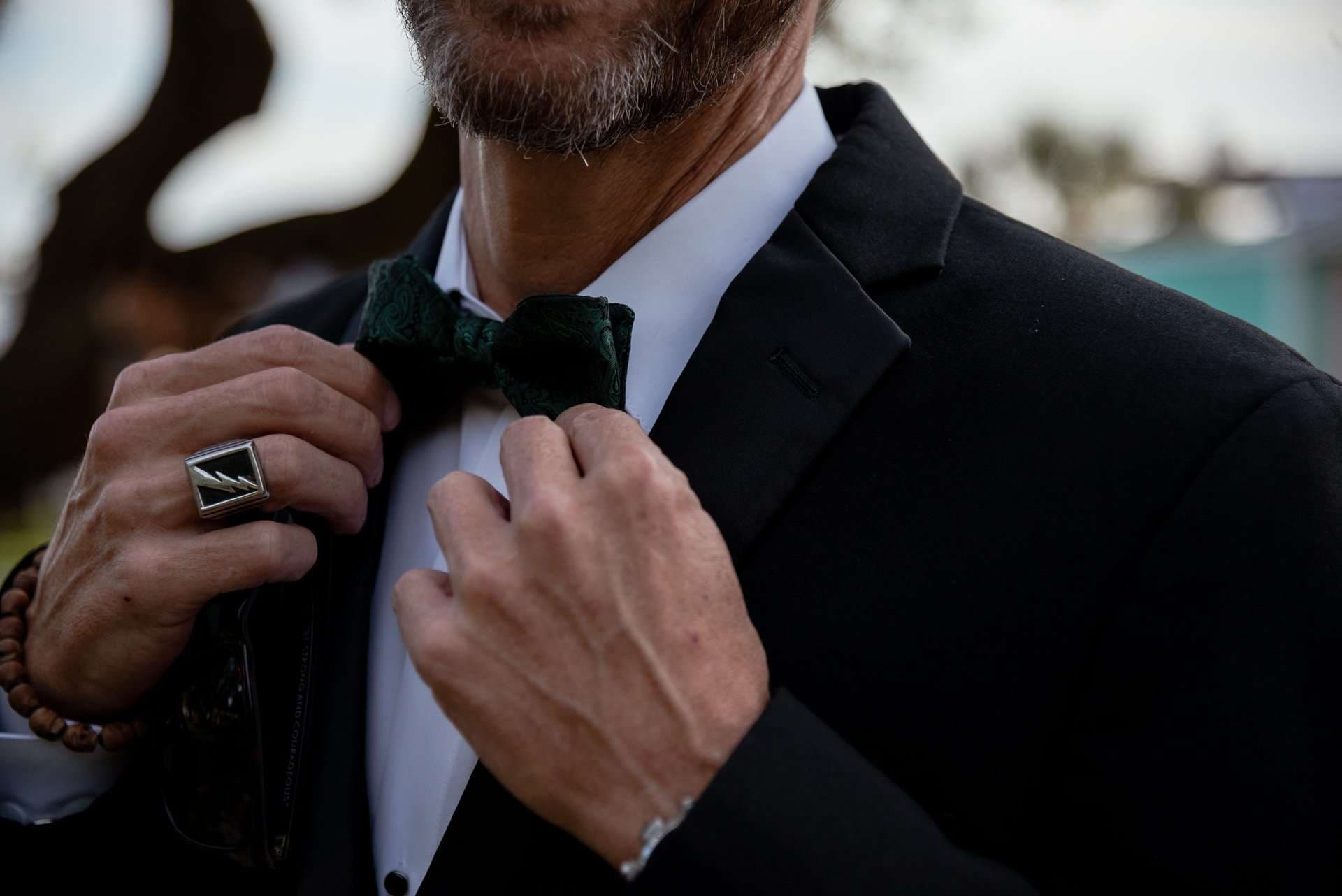 Man in a black suit adjusting a dark green velvet bow tie, wearing a silver ring and beaded bracelet