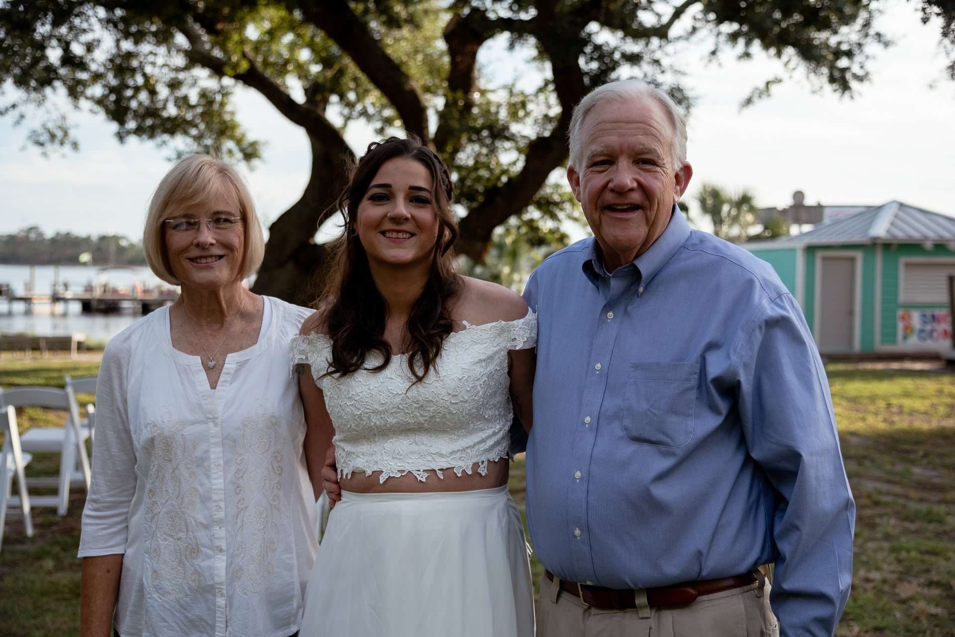 Smiling young woman in a white lace crop top and skirt posing between an older woman and an older man outdoors