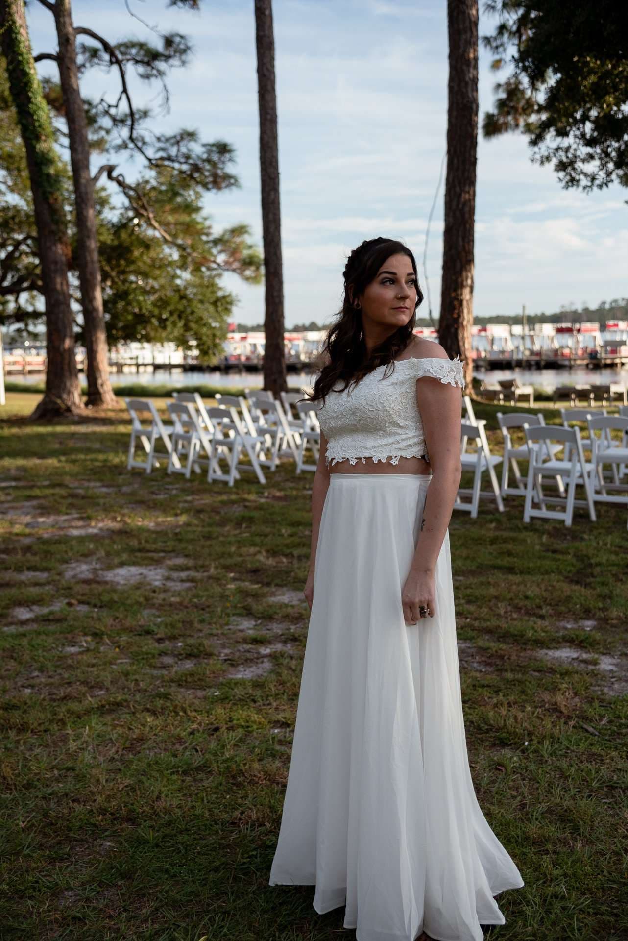 Woman in a white lace crop top and flowing skirt looking aside on a lawn with white chairs set up near the water