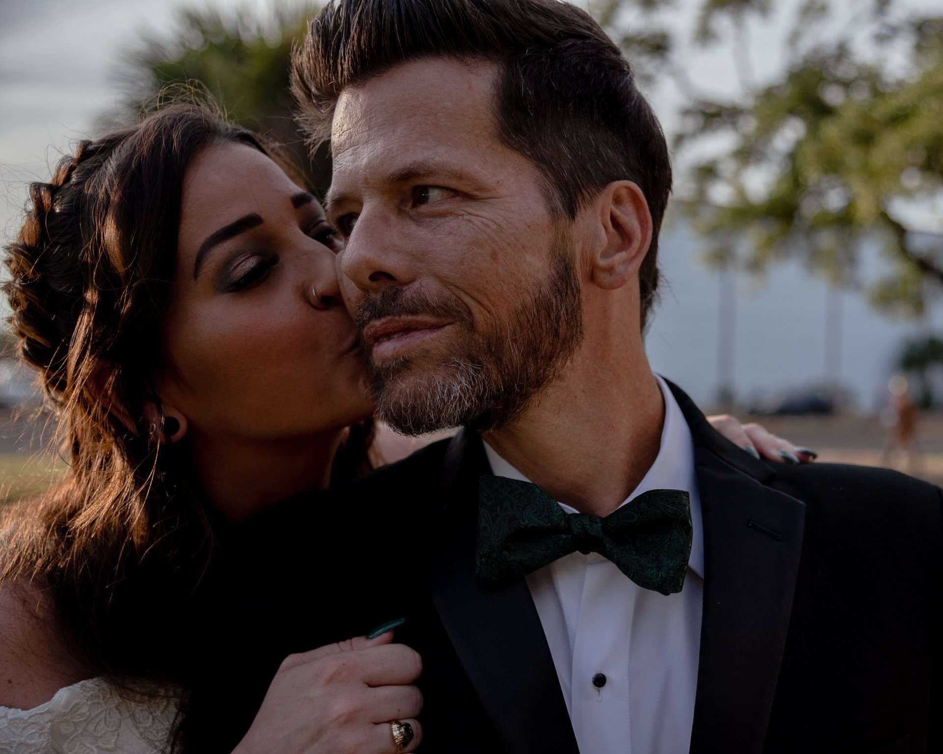 Bride in a white lace gown kissing the cheek of a bearded groom in a tuxedo and green velvet bow tie