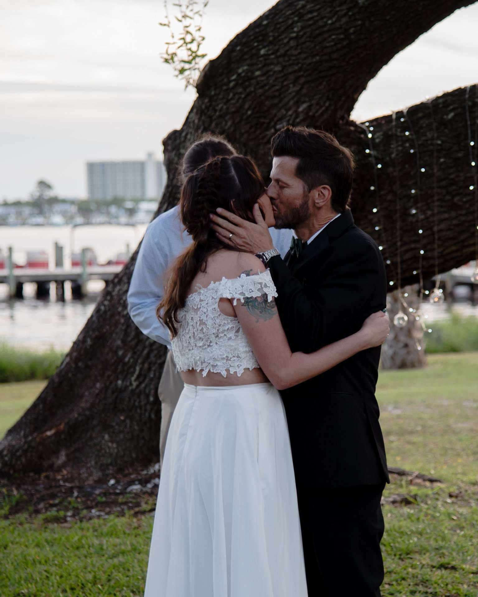 Couple kissing under a large oak tree draped with string lights, bride in a white lace crop top and skirt