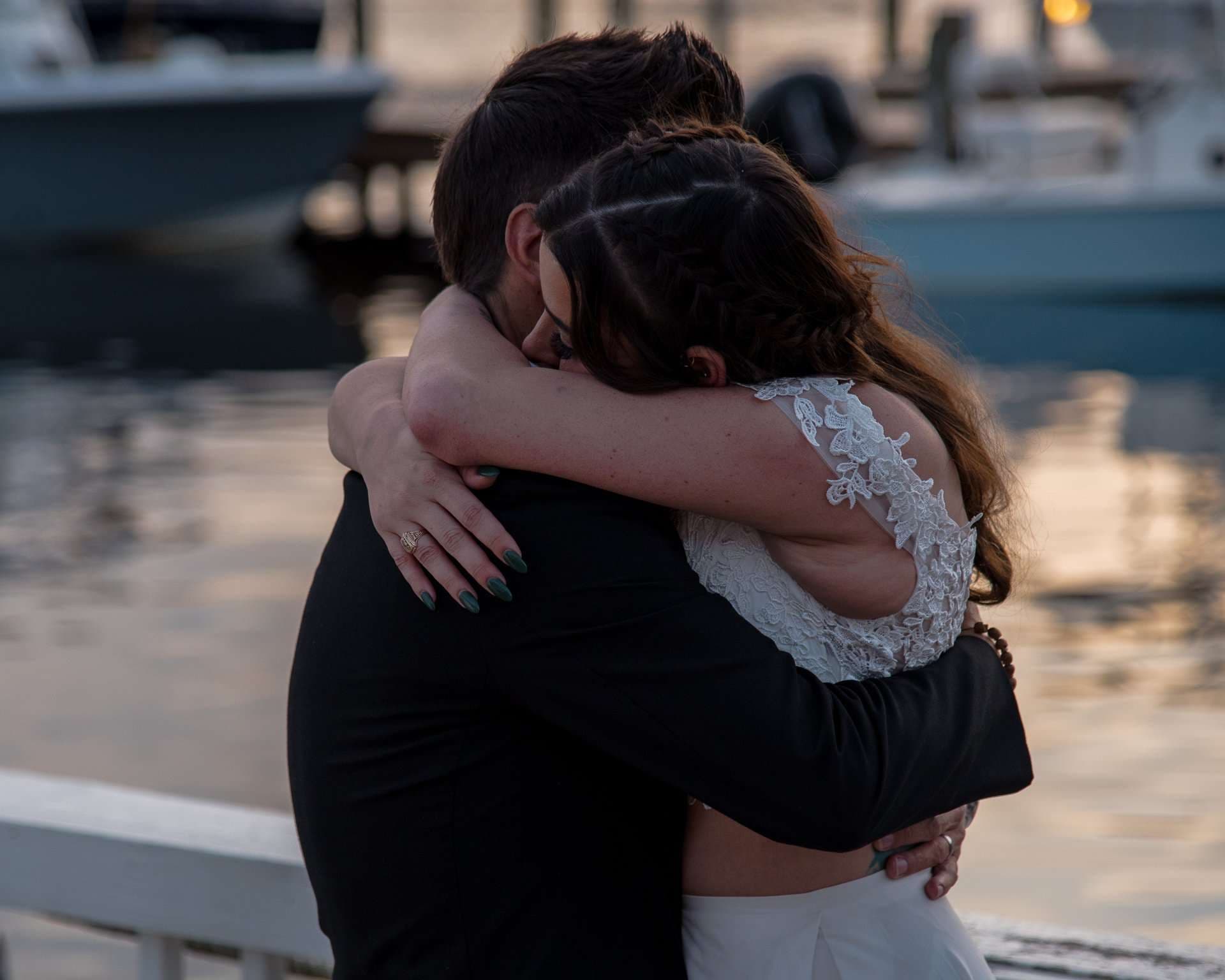 Couple embracing tightly on a waterfront dock at dusk, woman in a white lace top with braided hair, boats behind