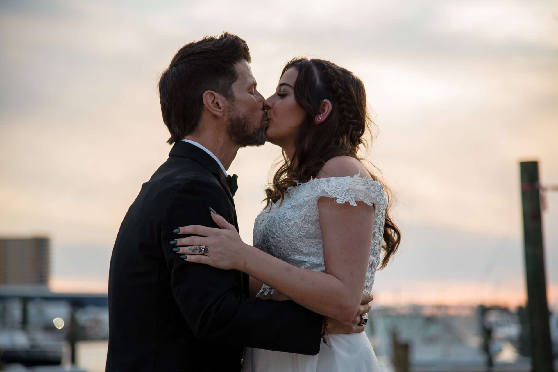 Couple in wedding attire kissing at sunset beside a marina, bride in a lace gown and groom in a black tuxedo