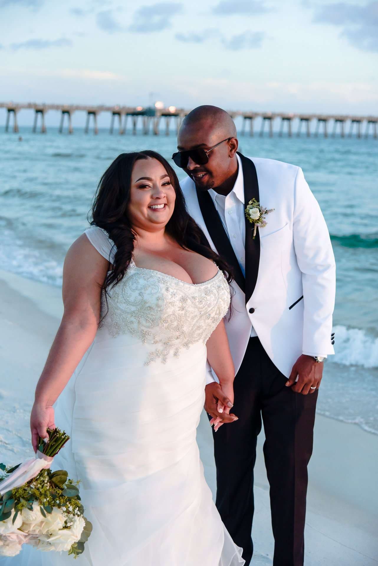 Smiling couple holding hands on the beach, bride in a beaded white gown and groom in a white tuxedo jacket