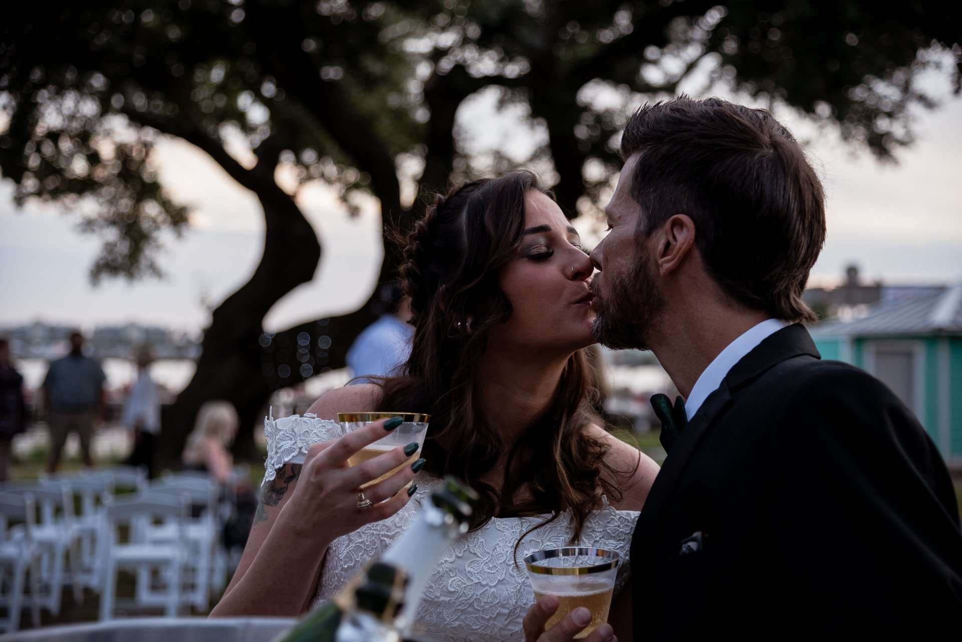 Couple kissing at an outdoor evening reception under an oak tree, holding gold-rimmed champagne glasses