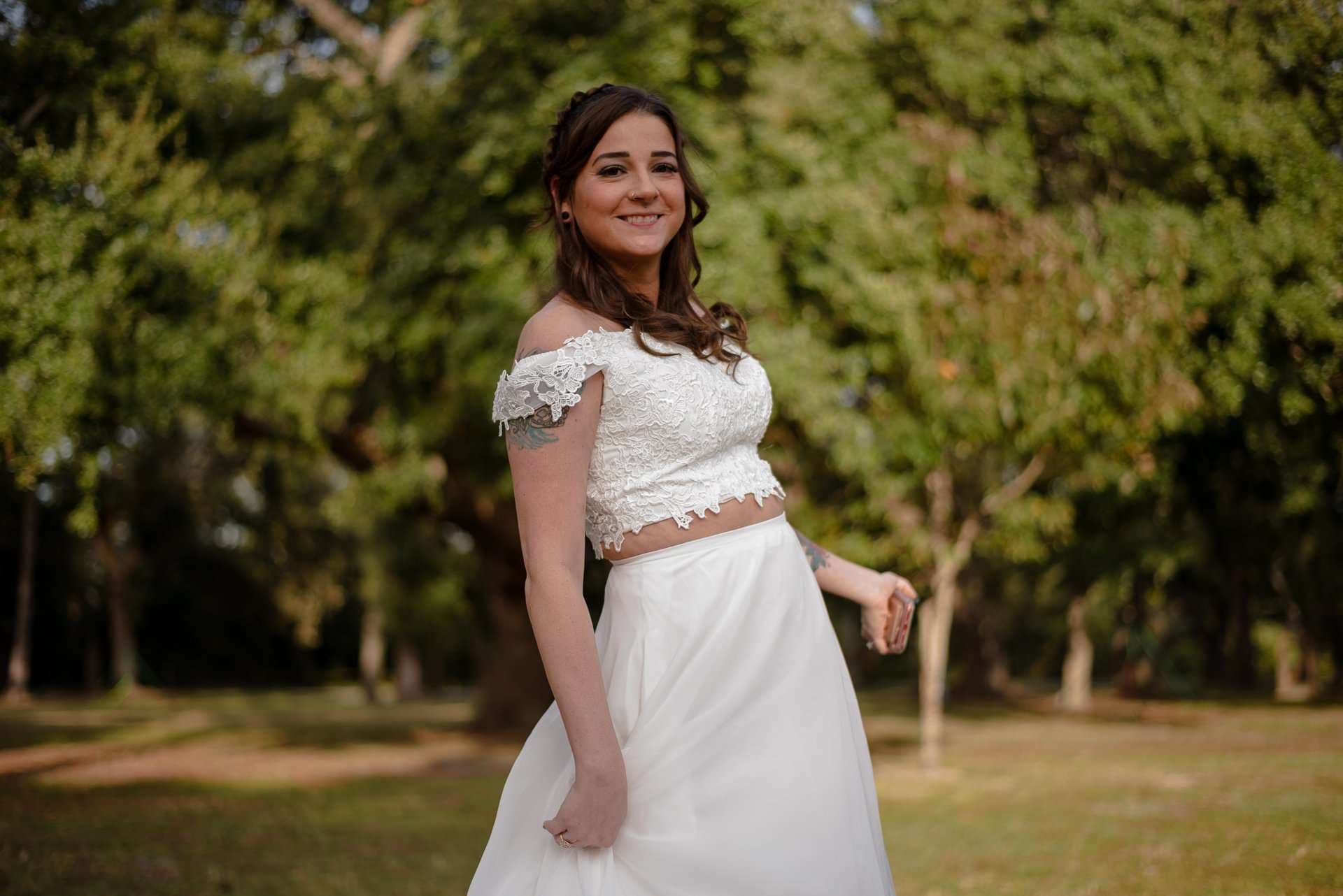 Smiling woman in a white lace off-shoulder crop top and flowing white skirt posing among sunlit trees in a park