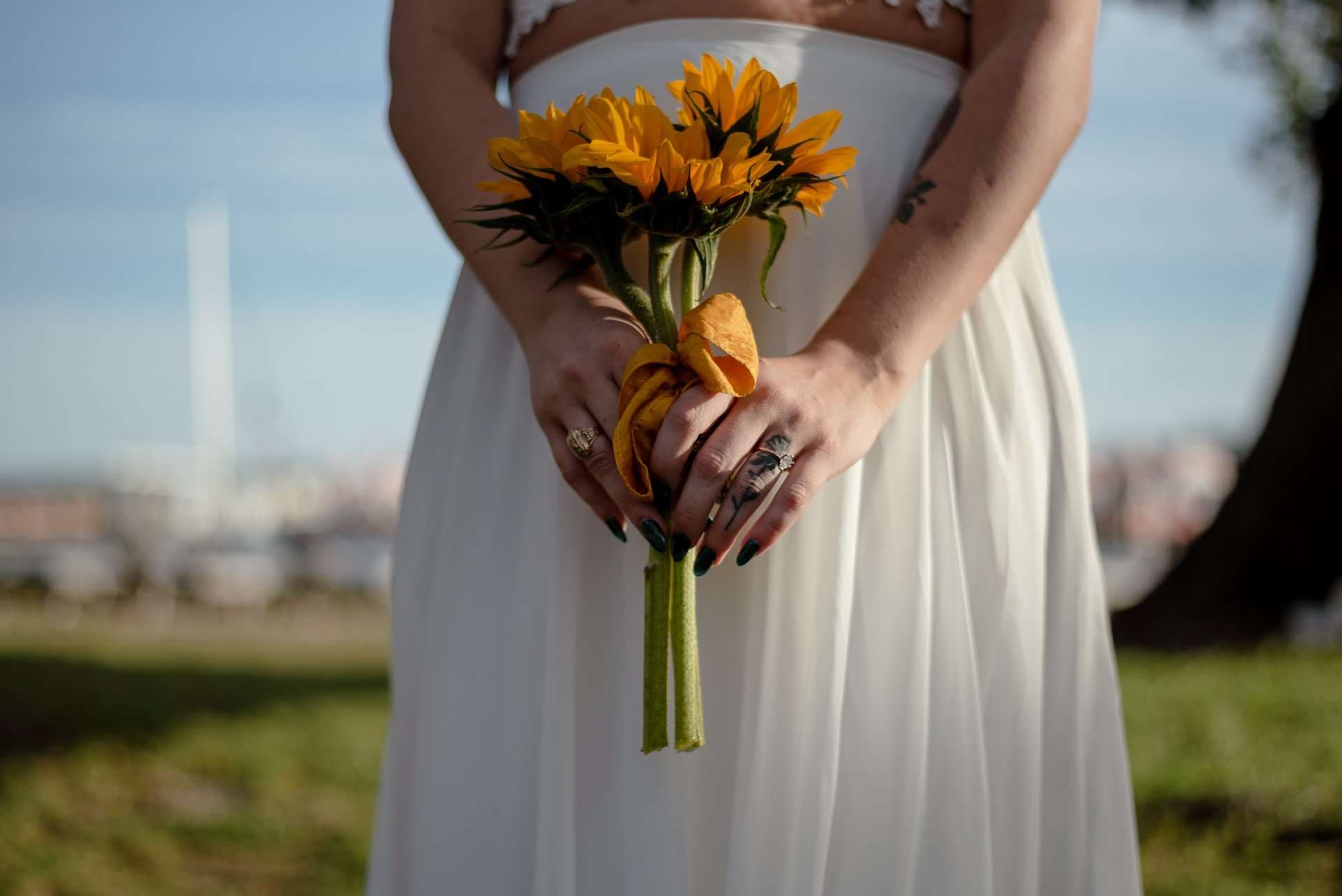Bride in a flowing white dress holding a small sunflower bouquet tied with a yellow ribbon on a grassy lawn