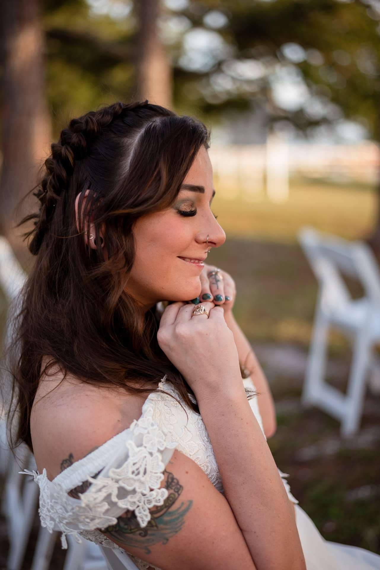 Smiling bride in a white lace gown with a braided half-up hairstyle and tattooed arm, hands raised to her chin outdoors