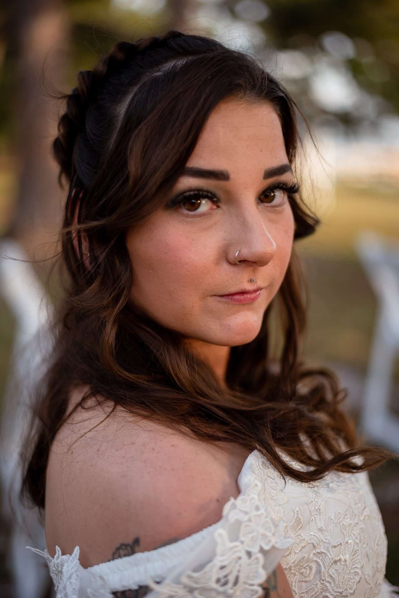 Woman in an off-the-shoulder white lace gown with braided half-up brown wavy hair glancing at the camera outdoors