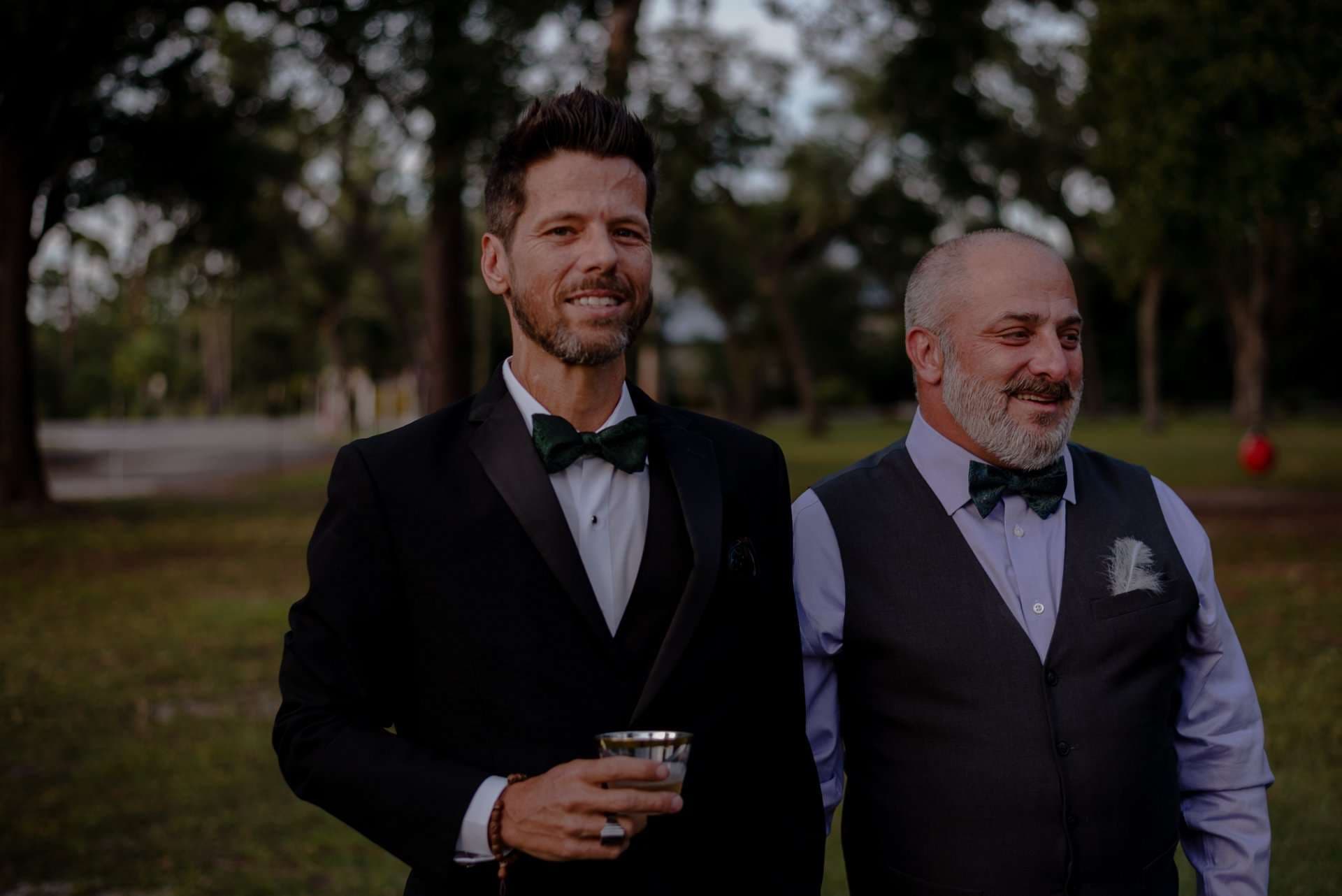 Two smiling men in bow ties, one in a black tuxedo holding a drink, standing together in a park at dusk