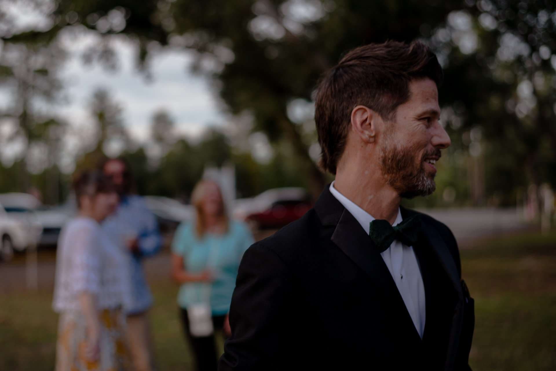 Smiling bearded man in a black tuxedo and green bow tie glancing sideways outdoors, guests blurred behind him