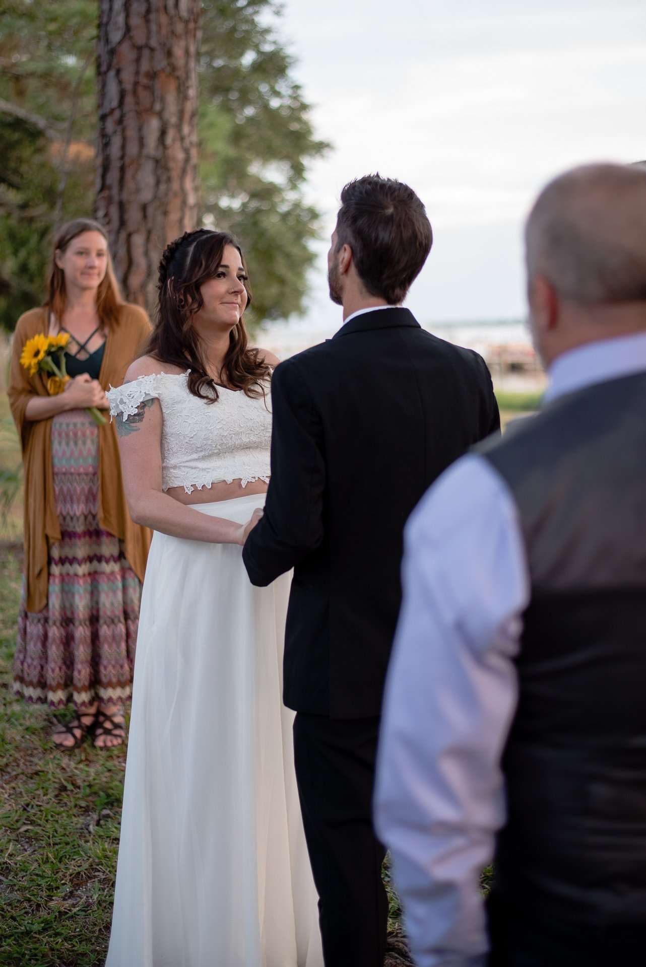 Smiling bride in a white lace crop top and skirt holds hands with her groom during an outdoor ceremony under pines