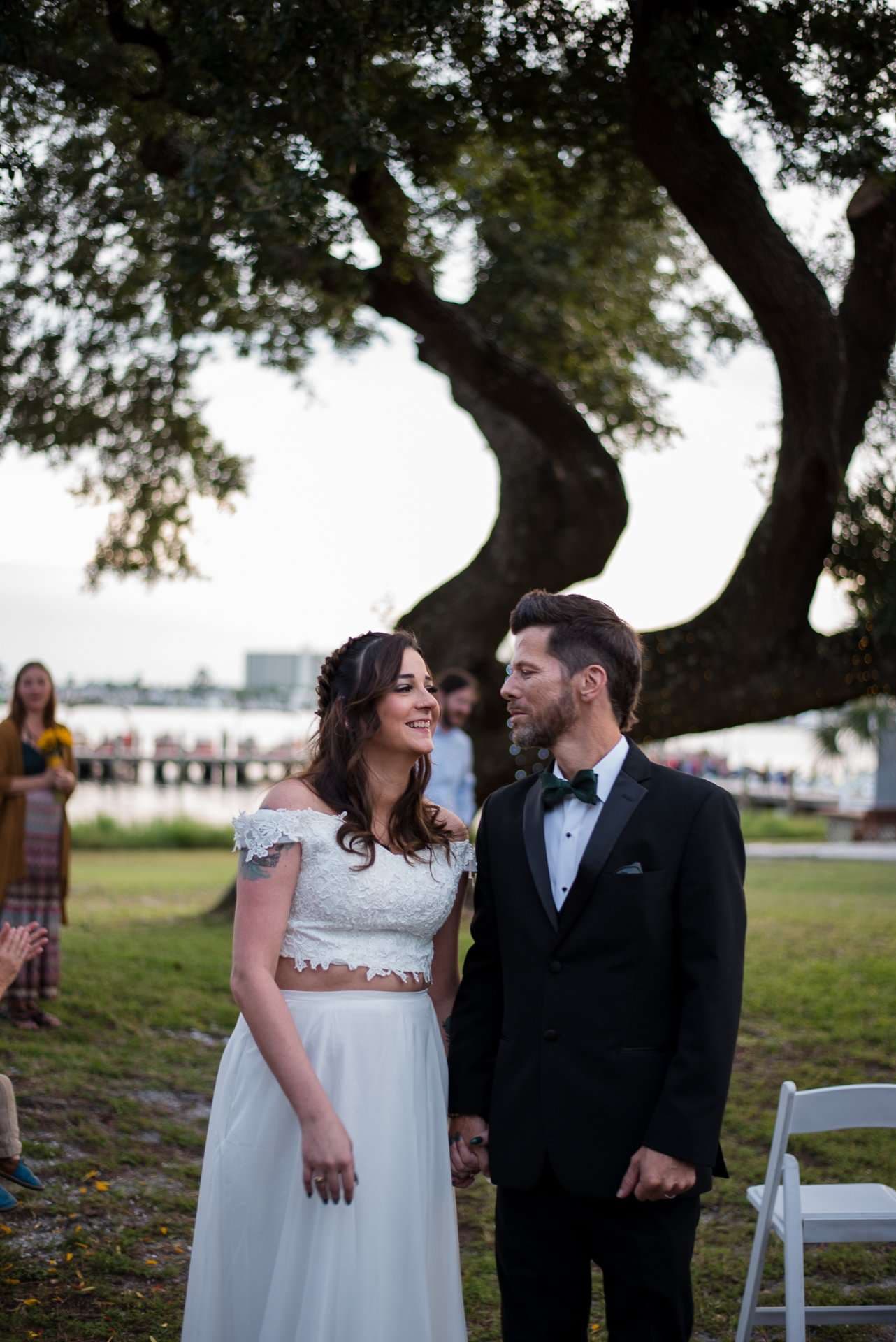 Couple holding hands and smiling at an outdoor ceremony under a large oak tree, bride in a white two-piece gown