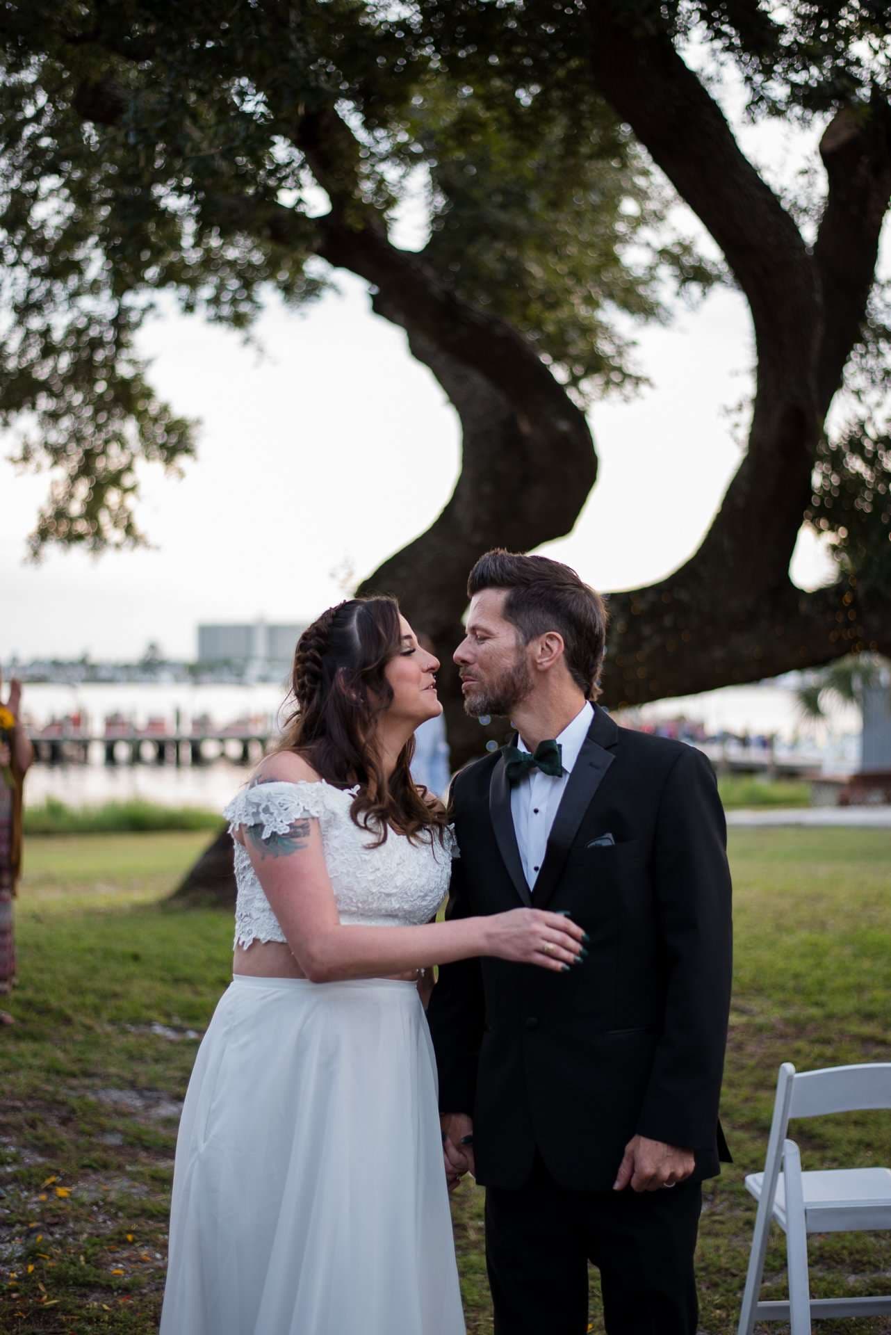 Couple about to kiss beneath a large oak tree on a waterfront lawn, she in a white lace top, he in a black tux