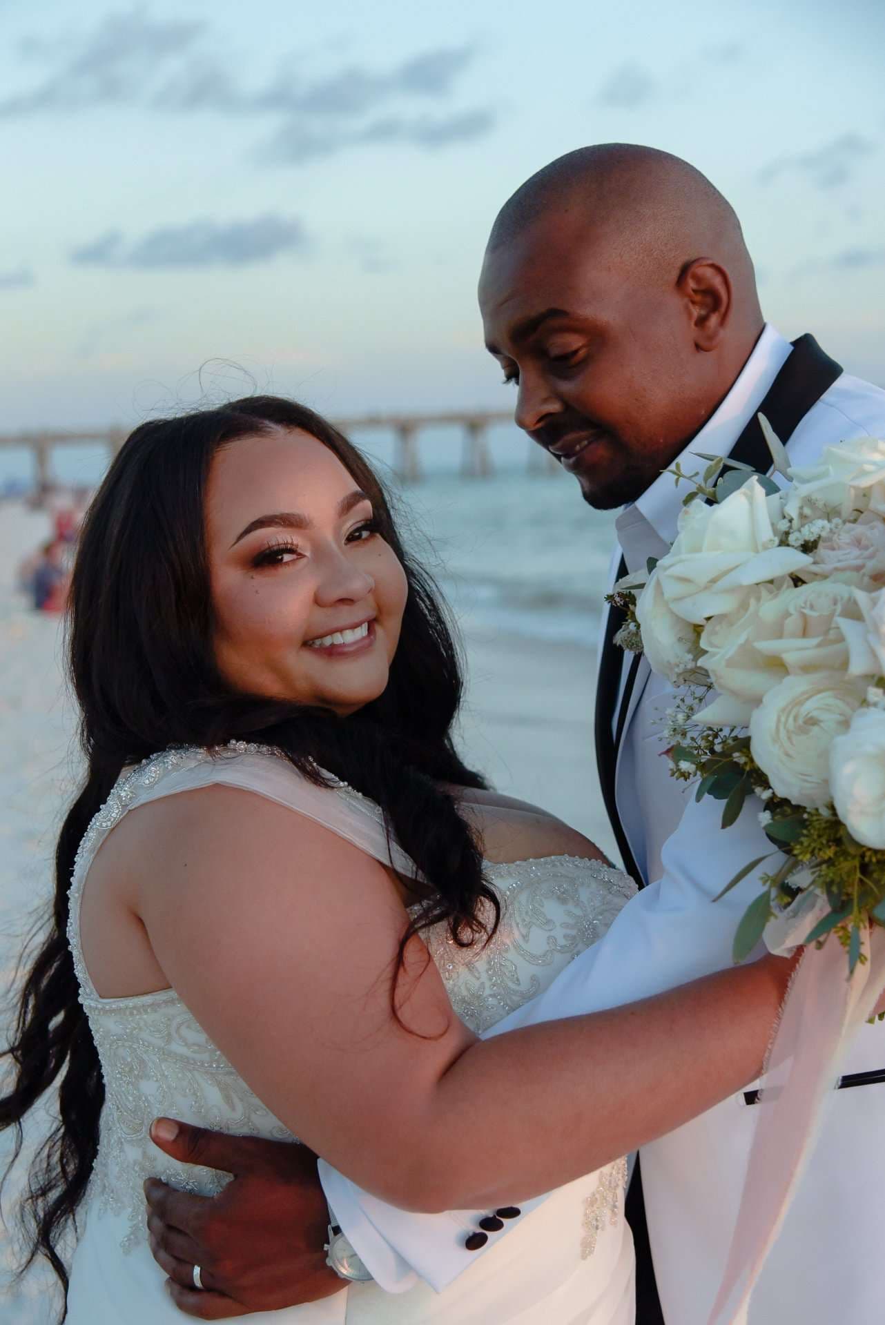 Smiling bride in a white lace gown embraced by her groom holding white roses on the beach at dusk