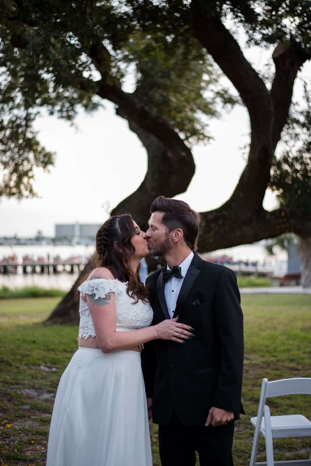 Couple kissing beneath a large oak tree by the water, she in a white lace two-piece, he in a black tuxedo