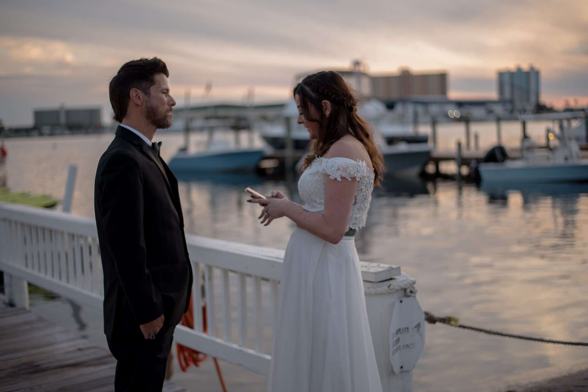 Couple facing each other on a white waterfront railing at sunset, the bride in a lace gown reading from a phone