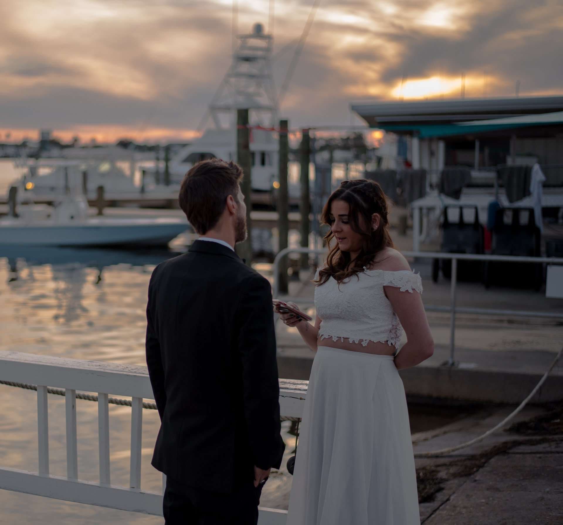 Couple in wedding attire stand facing each other on a marina dock at sunset, she reads from her phone