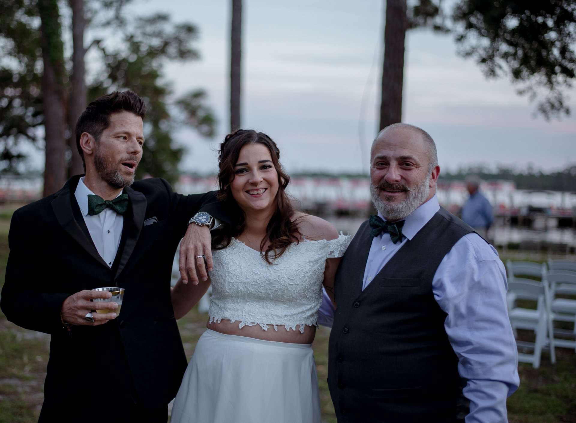 Smiling bride in a white lace top and skirt posing between two bearded men in bow ties at a lakeside dusk gathering