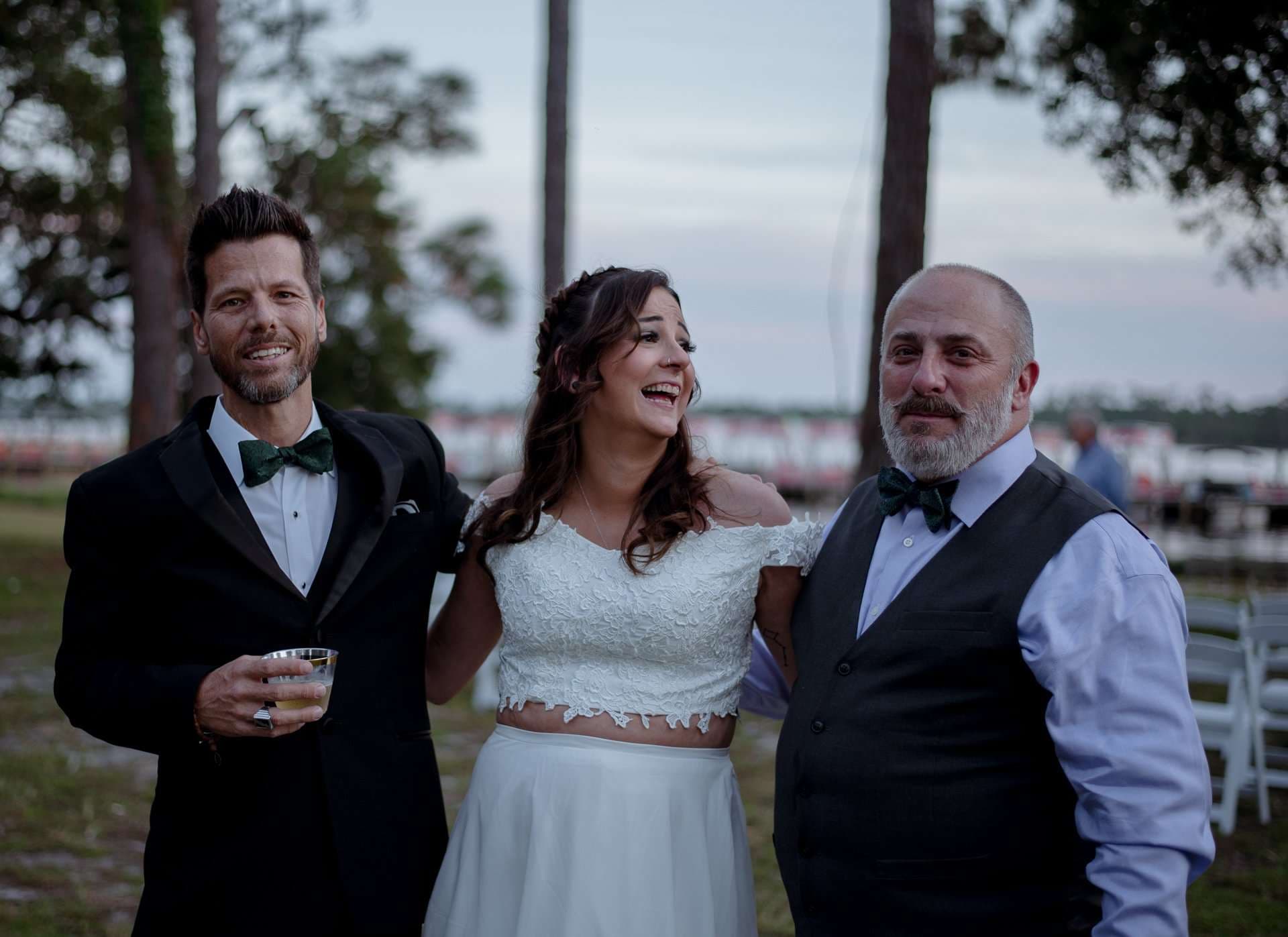 Laughing bride in a white lace top and skirt between two smiling men in bow ties at an outdoor evening gathering
