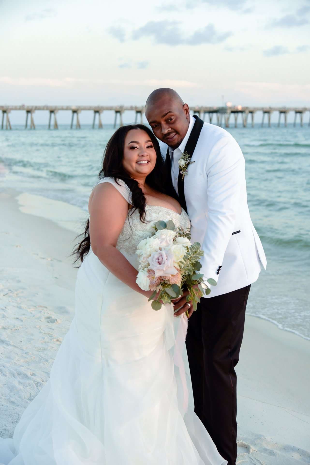 Couple embracing on the beach, bride in a white gown holding a bouquet, groom in a white tuxedo jacket, pier behind