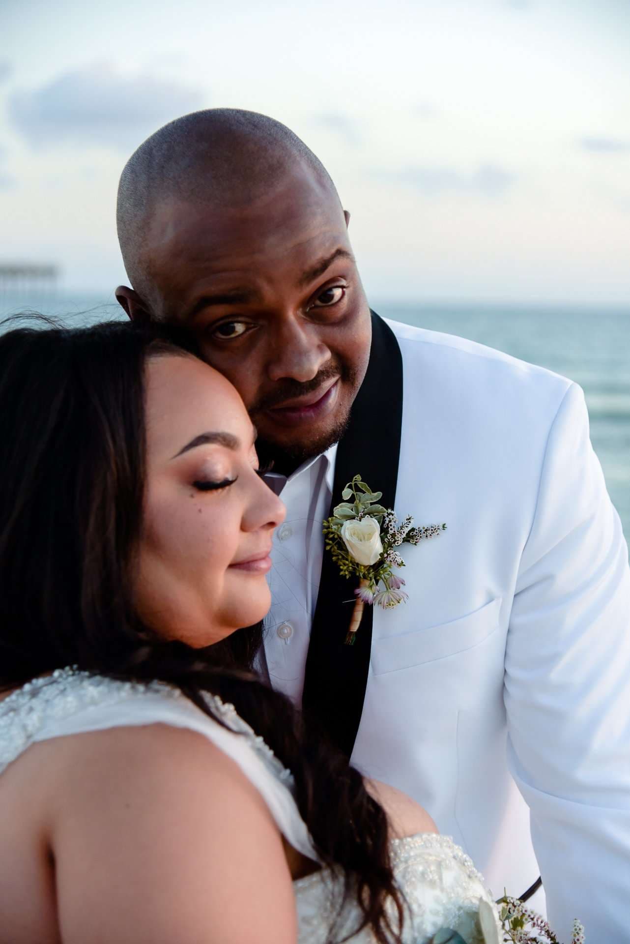 Couple embracing at the seaside, bride resting her head on the groom's chest, he wears a white tuxedo jacket with boutonniere