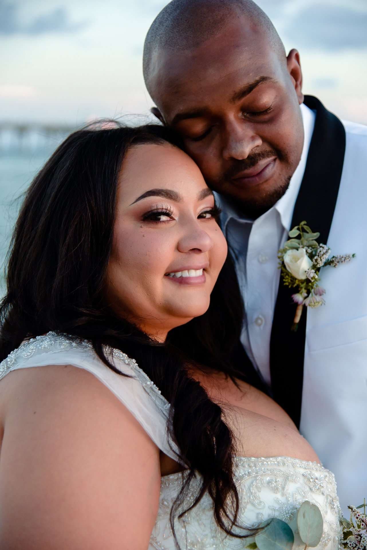 Smiling bride in a beaded white gown leaning into her groom, who rests his cheek on her head, eyes closed, by the water