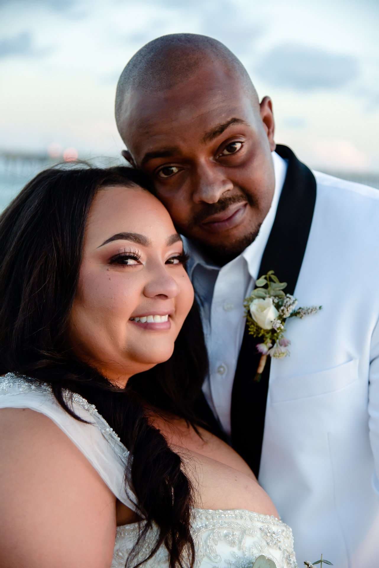 Smiling bride in a beaded white gown leaning against her groom, who wears a white jacket with a floral boutonniere