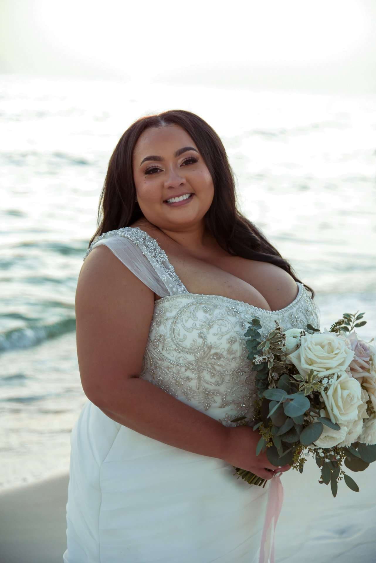 Smiling bride in a beaded white gown holding a white rose and eucalyptus bouquet on the beach by the water