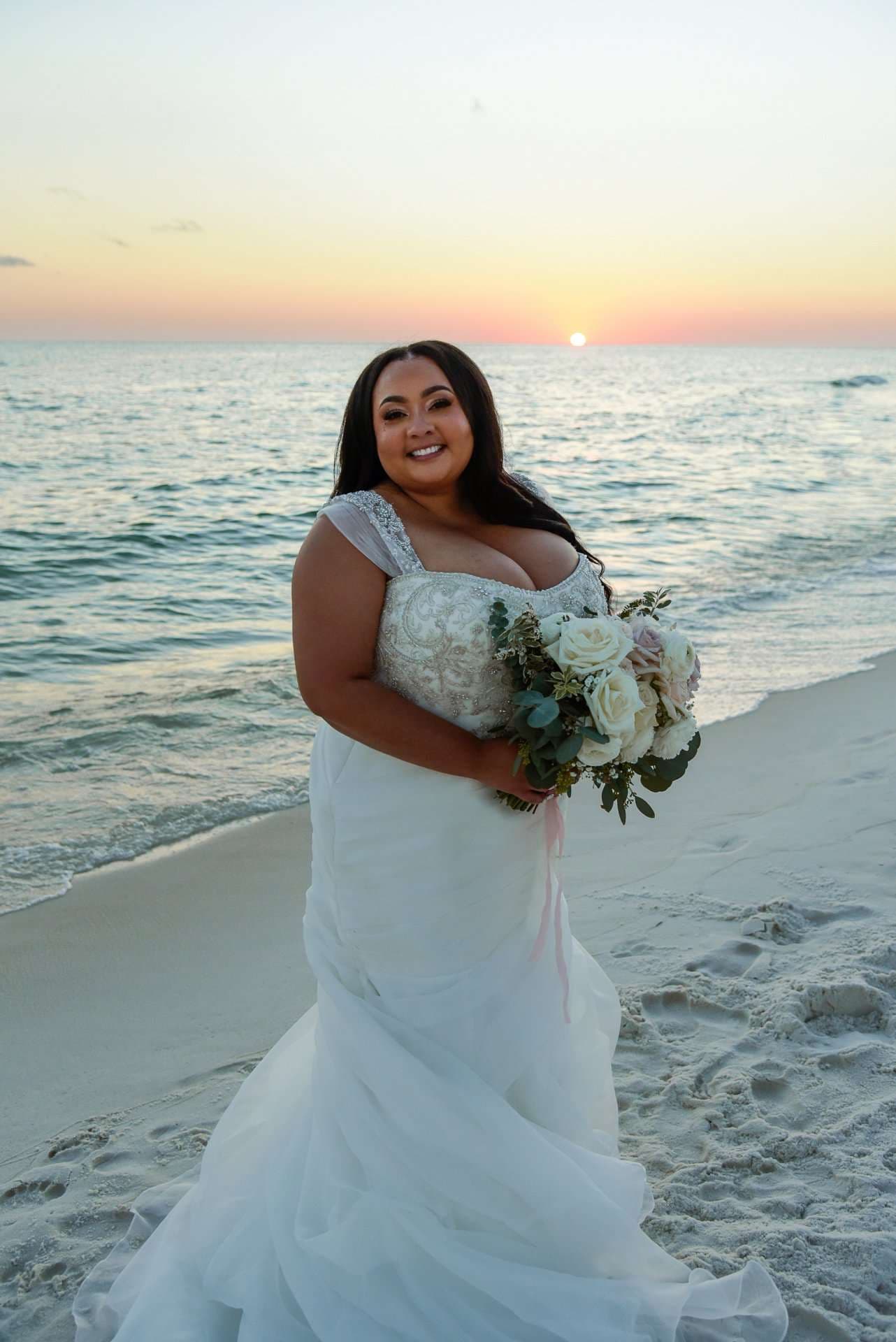 Smiling bride in a white lace and tulle gown holding a white rose bouquet on the beach at sunset