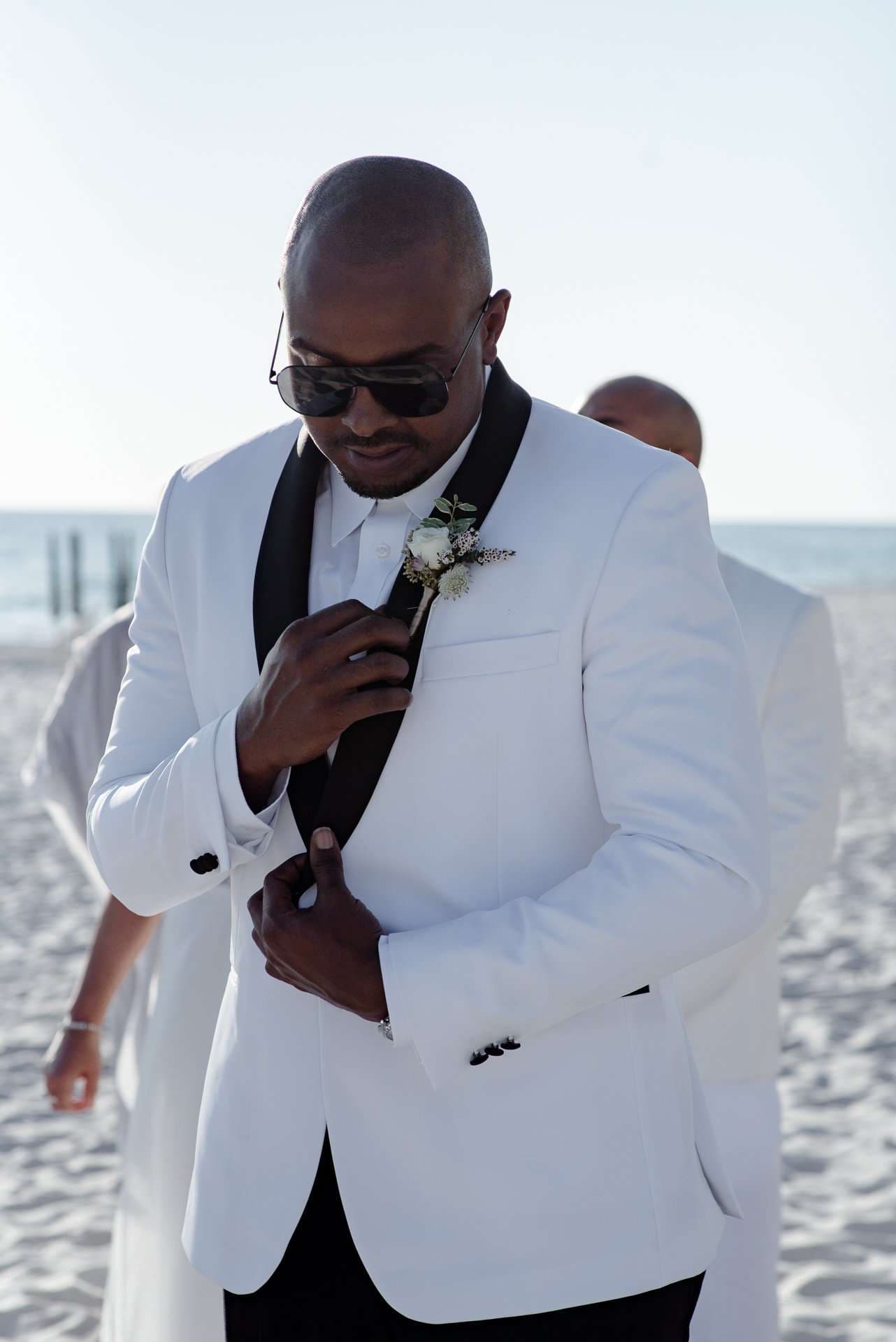 Man in a white tuxedo jacket with black lapels and sunglasses adjusts his boutonniere on a sunny beach