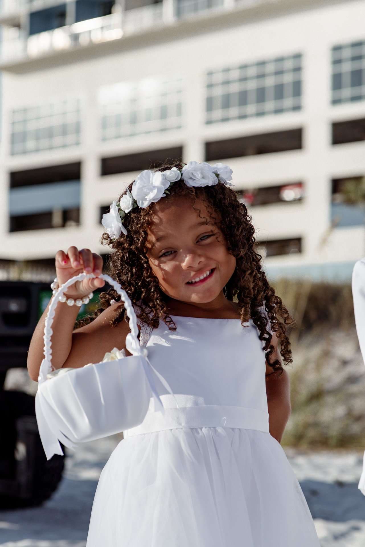Smiling flower girl with curly hair in a white dress and floral crown holding a white basket on the beach