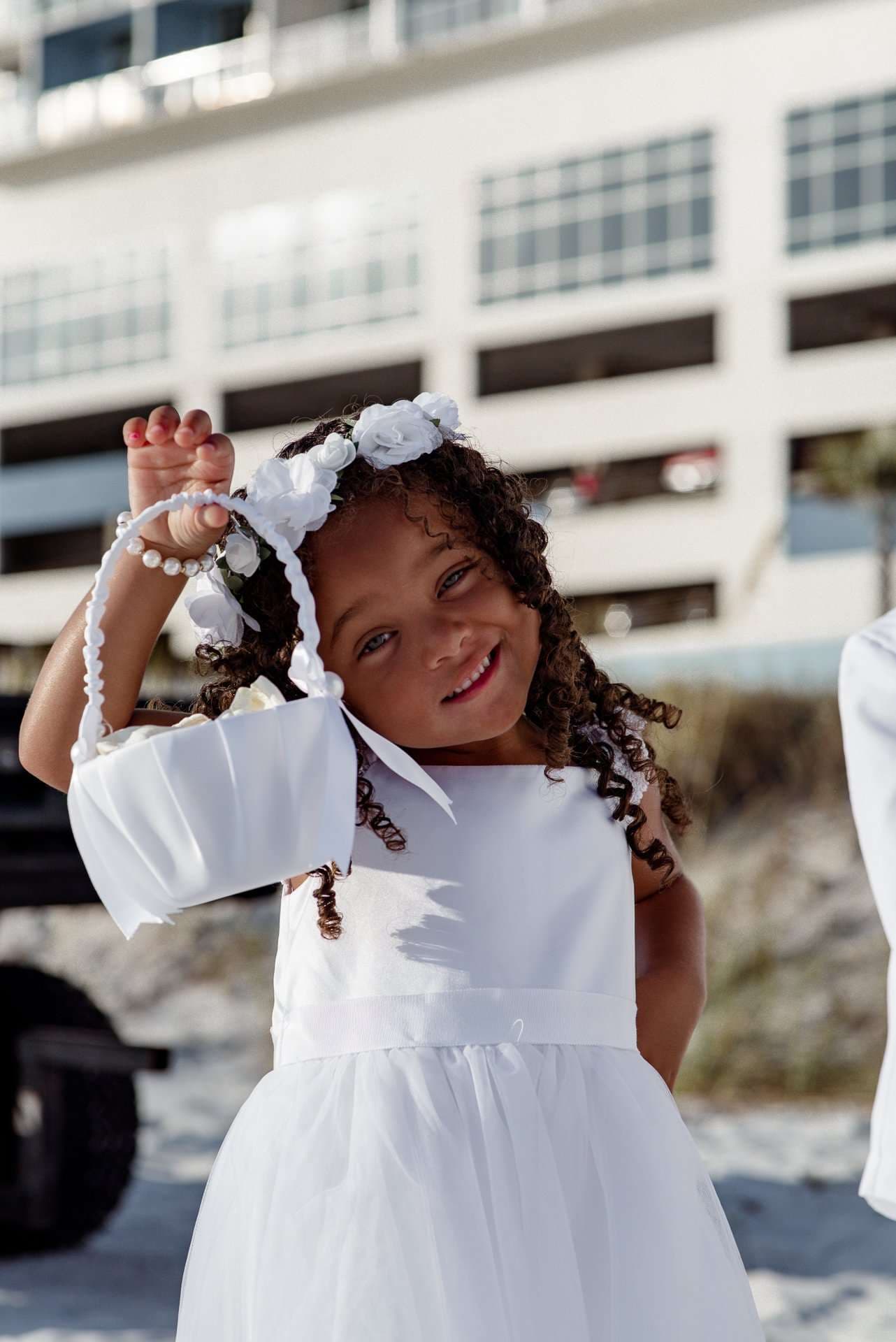 Smiling flower girl in a white dress and floral headpiece tilts her head while holding a white basket on the beach