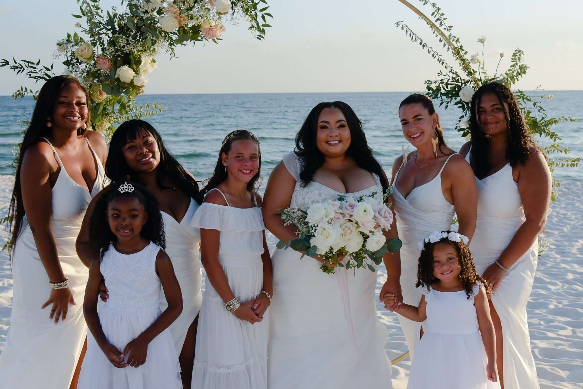 Bride holding a white rose bouquet, surrounded by bridesmaids and flower girls in white dresses on the beach