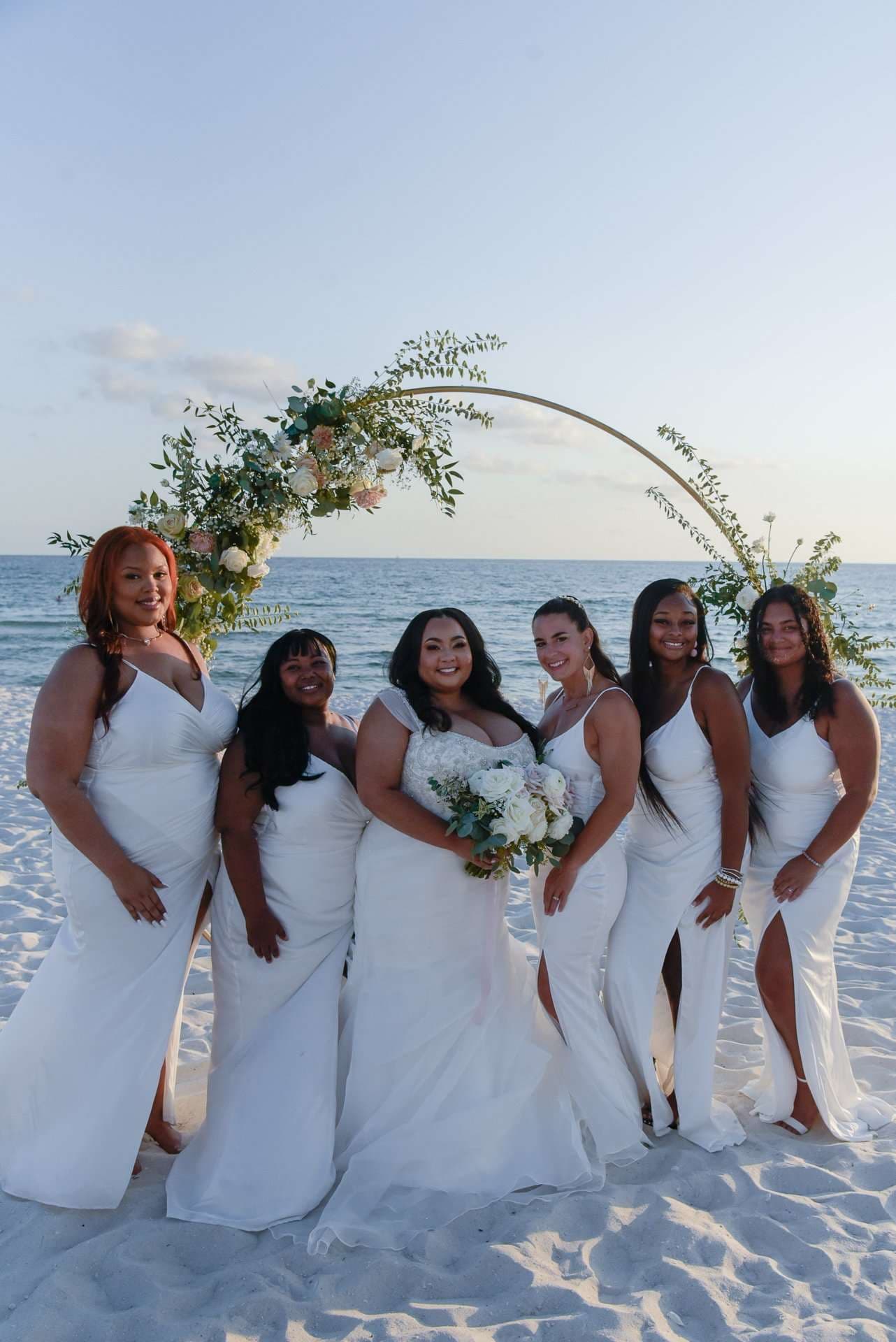 Bride holding a white bouquet, surrounded by five smiling women in white dresses under a floral arch on the beach