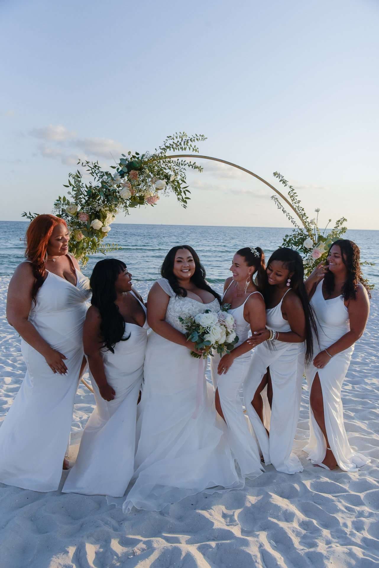 Bride holding a white bouquet laughs with four women in white dresses on the beach beneath a floral arch