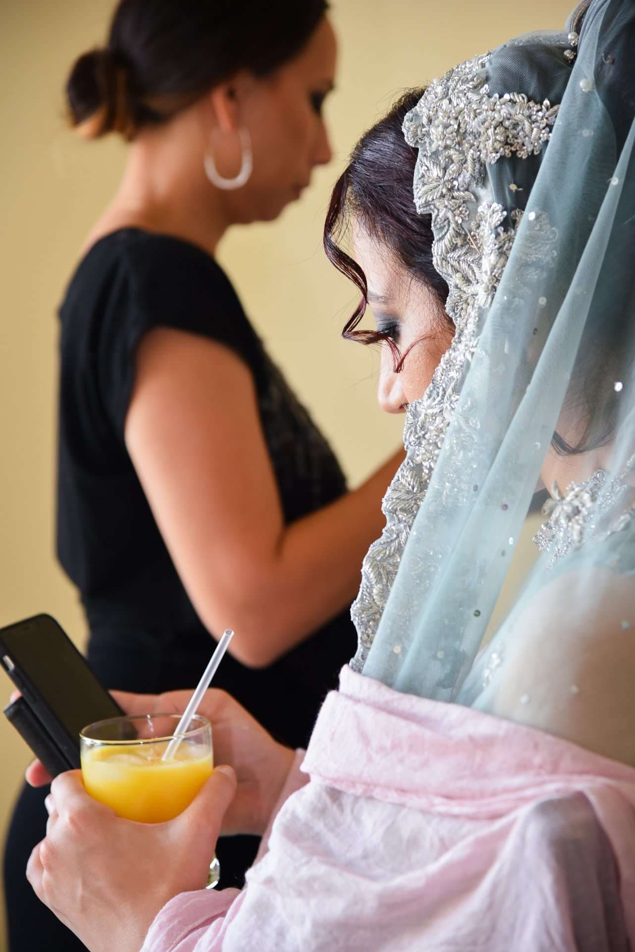 Bride in a beaded pale blue veil holds an orange juice and phone while a woman in black stands behind her