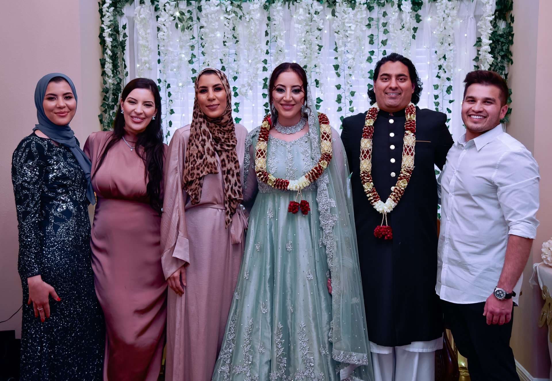 Bride in a sage green embroidered gown and groom in black, both wearing floral garlands, posing with four guests